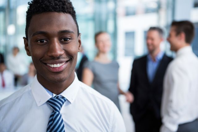 Young businessman smiling confidently in a modern office environment. Ideal for use in business-related content, corporate websites, career development materials, and professional networking platforms. Highlights themes of professionalism, confidence, and workplace success.