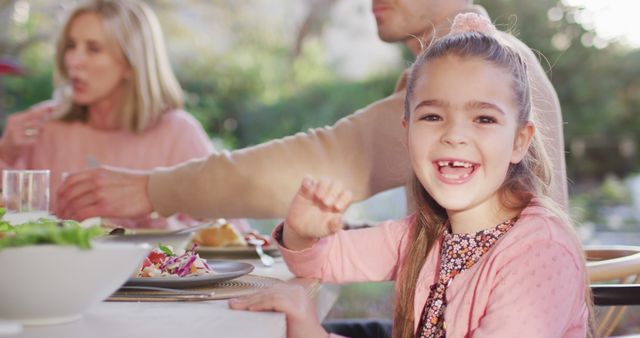 Young girl smiling at outdoor family dinner table, enjoying time with family. Suitable for family-themed advertisements, lifestyle blogs, and articles focused on family bonding and casual dining experiences.