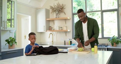 African american father preparing school lunch while son packing backpack in modern kitchen