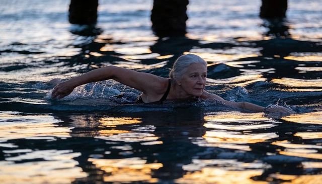 Senior woman swimming breaststroke at sunrise near pier pilings, calm ocean swim