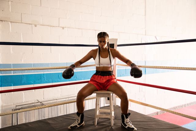Biracial female boxer practicing in a boxing gym wearing sports clothes, resting in a corner of boxing ring with boxing gloves on. Strength sports achievement.