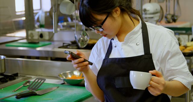 Young Asian female chef in professional kitchen looking at phone while holding a mug. Ideal for depicting modern cooking environments, culinary education, and technology integration in gastronomy. Useful for articles on modern kitchens, multitasking in culinary professions, and young professionals.