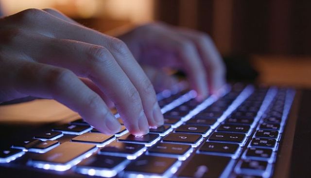 Pair of hands typing on backlit laptop keyboard in low light, glowing keys for remote work