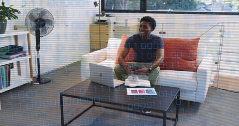 Young man playing video game on couch with laptop and controller in modern minimalist living room, d