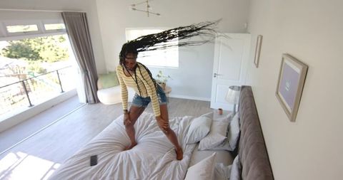 African american woman standing on bed with braids flying, sunlit airy bedroom lifestyle