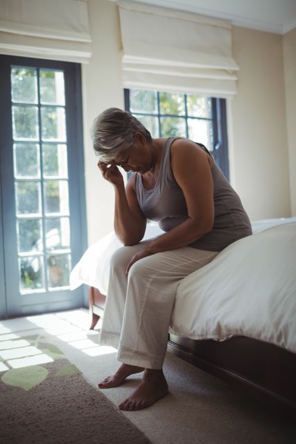 Senior woman sitting on bed, touching her head, appearing upset and stressed. Natural light from large windows illuminates the room. Useful for topics related to mental health, elderly care, loneliness, and emotional well-being.