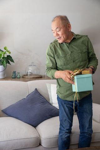 Senior asian man holding light blue gift box in cozy minimalist living room presenting surprise