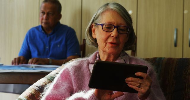 Senior woman using tablet while wearing glasses and pink sweater, with senior man reading newspaper in background. Useful for themes on senior lifestyle, technology usage among elderly, leisure activities, and comfortable home settings.