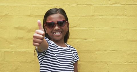 Happy girl with heart sunglasses giving thumbs up against yellow wall