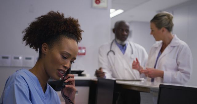 Biracial hospital worker in reception talking on phone with doctors in backgrund - Download Free Stock Photos Pikwizard.com