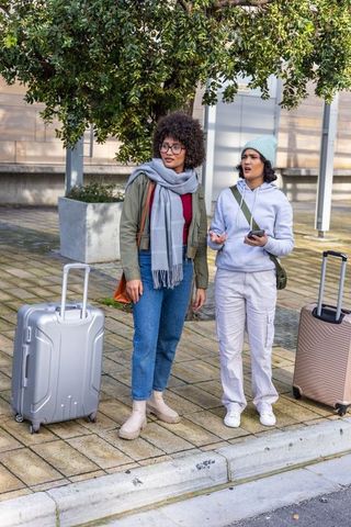 Female friends standing at curb with rolling luggage and smartphone urban travel moment