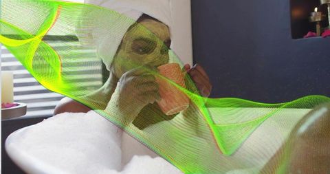 Black woman relaxing in tub with green facial mask and towel turban sipping coral mug