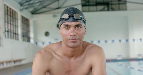 Competitive swimmer preparing at pool edge wearing black cap and goggles, indoor portrait