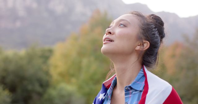 A young woman wrapped in an American flag is outdoors surrounded by greenery and mountains. She looks up with a smile, portraying a sense of pride and contemplation. Ideal for use in themes related to patriotism, national holidays like 4th of July, and celebrations of independence and freedom. Works well in promotional materials for government campaigns, outdoor adventure brands, and community initiatives highlighting diversity and American values.