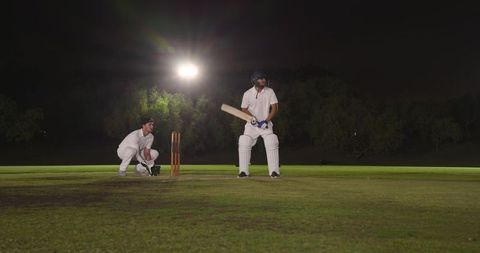 Night cricket action with batsman and wicketkeeper under floodlights on grass pitch