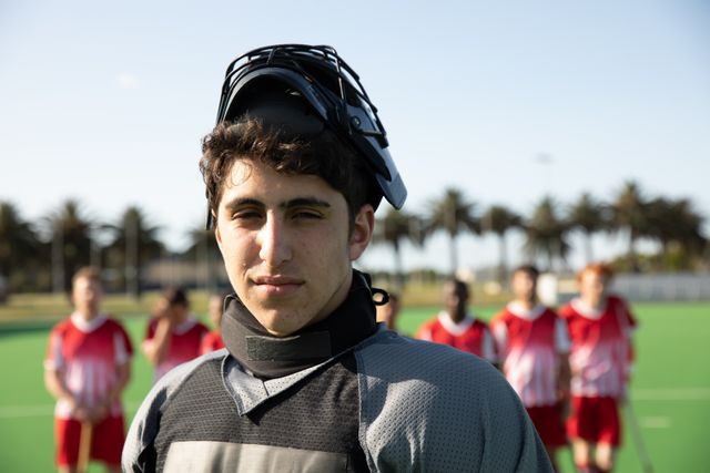Portrait of Caucasian male field hockey goalkeeper, wearing black hockey uniform and a helmet, standing on the pitch and looking to camera, with his teammates in background. Sport game competition.