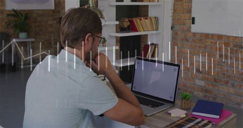 Contemplative man thinking at laptop in modern brick office workspace with notebooks