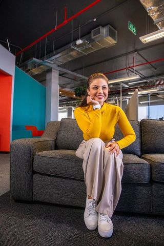 Smiling woman sitting on grey sofa in modern open-plan office lounge wearing mustard top
