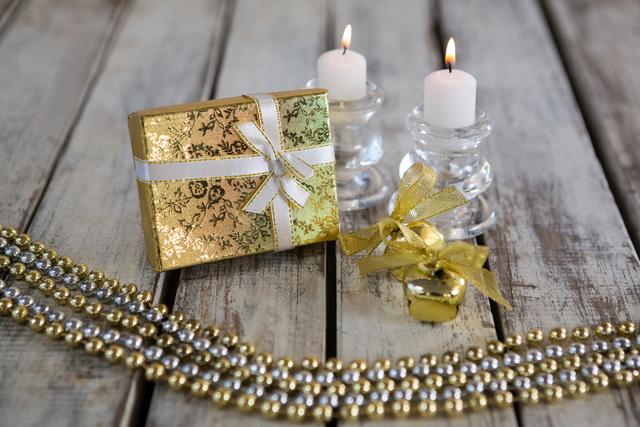 Christmas gift wrapped in gold paper with white ribbon, accompanied by lit candles and gold beads on rustic wooden plank. Ideal for holiday greeting cards, festive advertisements, seasonal blog posts, and Christmas-themed social media content.