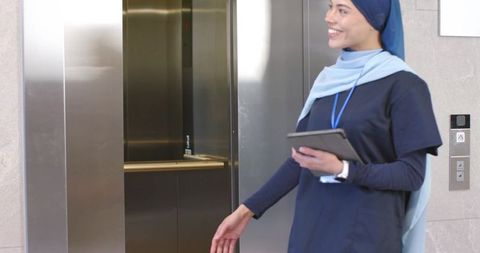 Female healthcare worker wearing hijab holding tablet by open elevator in hospital lobby