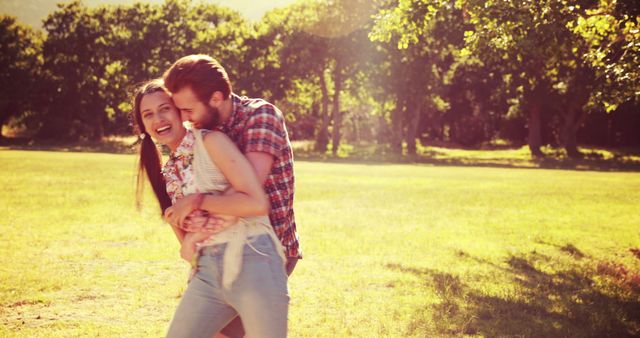 Couple enjoying a playful moment in the sunlit park. They are laughing and embracing in a lush green park under the sun. Ideal for illustrating themes of love, happiness, romance, outdoor activities, young couples, positive emotions, and summer leisure activities.