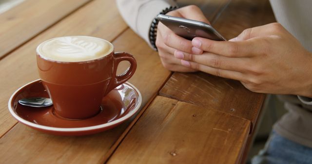 Person sitting in café, texting on smartphone while enjoying a latte. Ideal for use in articles, blogs, or advertisements about café culture, technology in daily life, relaxation, and morning routines.