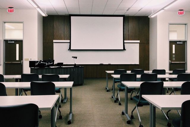 Image depicts an empty classroom with rows of desks and chairs, a ceiling-mounted projector, and a large whiteboard in front. Good for depicting educational environments, academic settings, modern classrooms, training seminars, learning spaces, and school infrastructure.