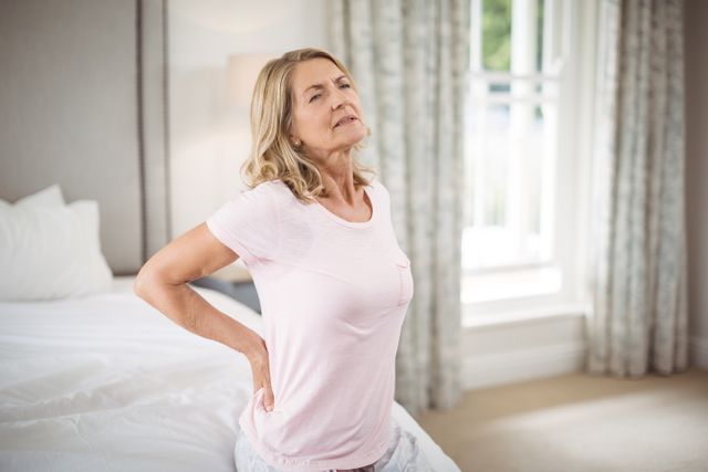 Senior woman holding her back in pain, standing beside bed in cozy bedroom. Ideal for articles related to senior health, back pain treatments, aging concerns, or home care for elderly. Useful for medical, health, and lifestyle publications.