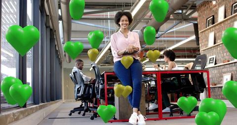 Smiling professional holding tablet leaning on desk in modern coworking with green heart balloons