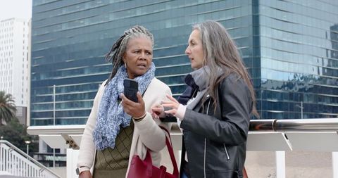 Mature women having conversation on urban bridge holding smartphone and red tote near glass facade