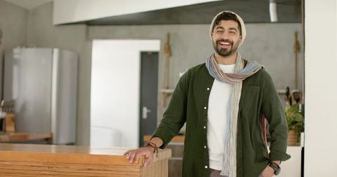 Indian man leaning on wooden kitchen counter smiling in minimalist open-plan concrete home