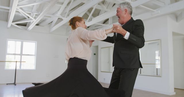 Senior couple dancing enthusiastically in a well-lit dance studio. Both are dressed in elegant attire with the man in a suit and the woman in a flowing skirt. This image can be used for promoting active senior lifestyles, dance classes, ballroom dancing events, or senior community activities. Ideal for websites, brochures, and advertising focused on dance, fitness, and senior engagement.