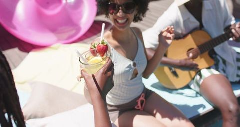 Laughing african friends sipping cocktails while playing guitar on sunny pool deck