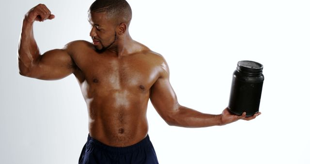 An African American man exhibits his muscular physique while holding a supplement container, with copy space. His strong build and confident pose suggest a focus on fitness and health.
