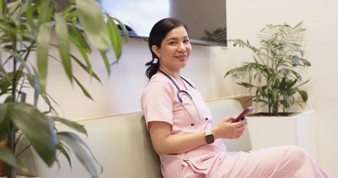 Smiling nurse in pink scrubs sitting in modern clinic lobby holding smartphone and stethoscope