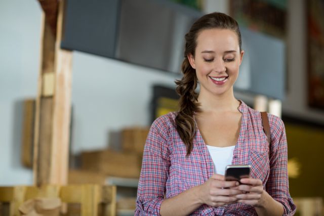 Young woman in casual plaid shirt smiling while using mobile phone in grocery store. Ideal for illustrating concepts of modern shopping, customer satisfaction, technology in retail, and everyday lifestyle.