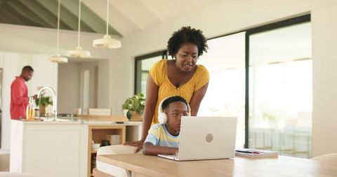 African american mother guiding son on laptop for online learning in sunlit kitchen