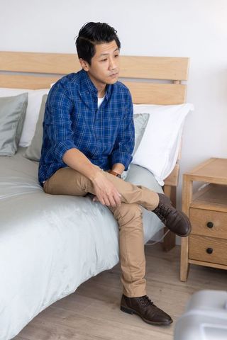 Asian man sitting on bed edge with suitcase in modern bedroom looking thoughtful
