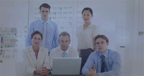 Corporate leadership team posing around laptop in modern glass office with city skyline