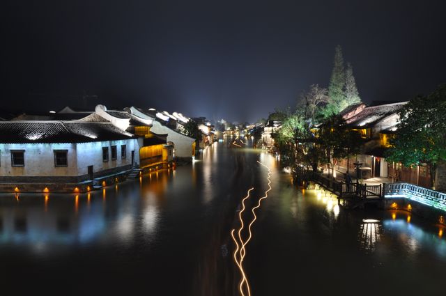 This image features a tranquil nighttime scene of a traditional Chinese water town with beautifully illuminated buildings reflecting on calm waters. The long exposure lights create flowing patterns, adding an artistic touch. Ideal for travel blogs, tourism promotions, cultural heritage features, or serene nightscape collections.