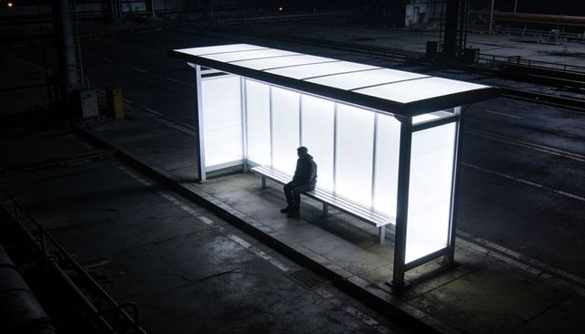 Lonely hooded figure sitting under backlit shelter at night on deserted urban platform