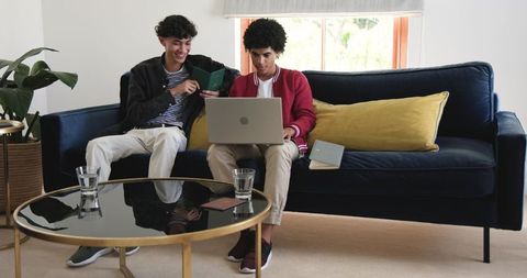 Two friends sharing laptop on navy sofa in modern living room, collaborating on project
