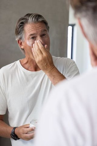Mature man applying facial moisturizer at bathroom mirror practicing daily skincare routine