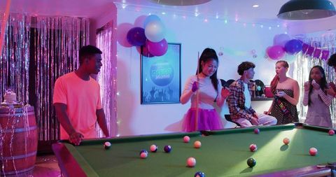 Teen friends playing pool and celebrating neon birthday party in retro game room