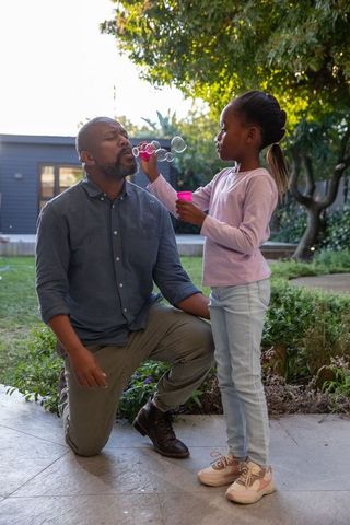 African american father kneeling while daughter blowing bubbles in sunlit backyard garden