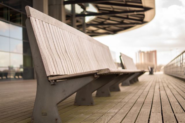 Empty wooden benches aligned in urban outdoor public space. Modern architecture forms backdrop, reflecting contemporary design. Ideal for use in settings highlighting urban leisure areas, public facilities design, or civic planning.