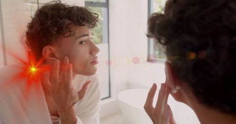 Young man inspecting ear in mirror wearing bathrobe sunlit bathroom with lens flare