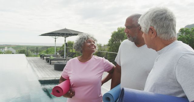 Senior friends gather near an outdoor pool, preparing for yoga. They are holding mats and smiling, dressed in casual athletic wear. Perfect for depicting active aging, group fitness activities, healthy living, and outdoor wellness practices. Great for wellness blogs, senior lifestyle magazines, fitness program promotions, and advertisements focusing on longevity and group activities.