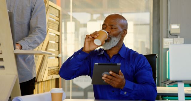 Mature man in formal blue shirt enjoying coffee while using a digital tablet in a modern office. Colleague working on a standing desk in background. Ideal for promoting workplace productivity, modern work environments, business technology usage, and daily workspace routines. Useful for websites and articles focused on office culture, professional lifestyle, and balanced work life.