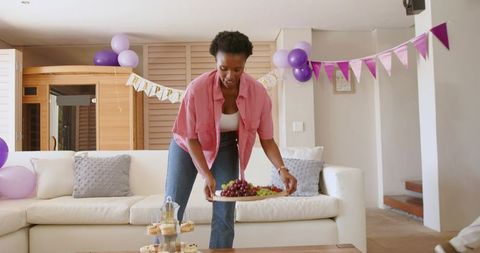 African american woman placing fruit platter in living room party with purple bunting